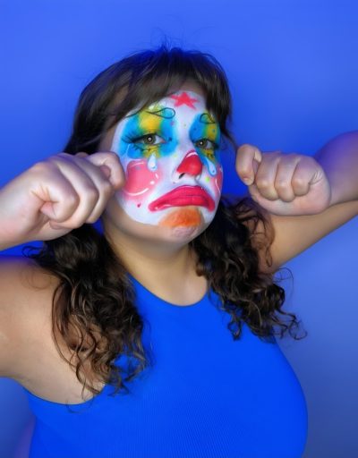 Modelo frotándose los ojos con los puños en gesto de llanto, maquillaje de payaso triste