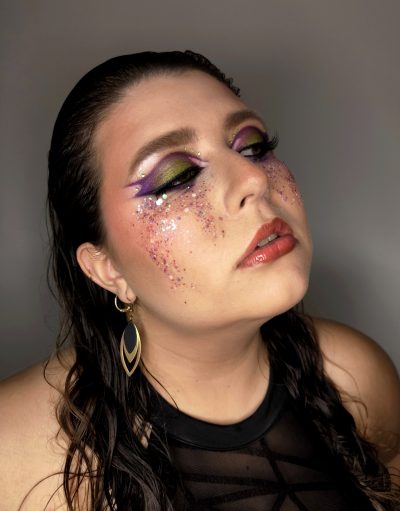 Maquillaje creativo full face con sombras moradas y verdes, delineado marcado, pedrería y purpurina rosa y plateada.
