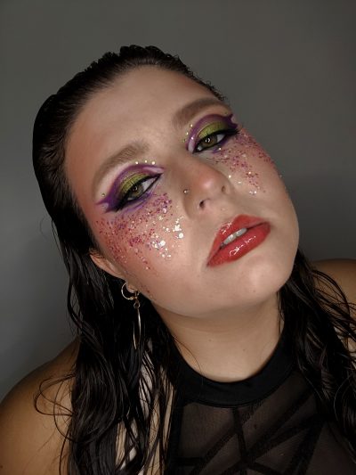 Detalle del maquillaje glam con delineado morado alado, sombra verde, pedrería y purpurina bajo los ojos.