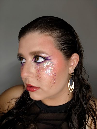 Perfil de un maquillaje glam con delineado morado, purpurina en la mejilla y pendientes dorados.